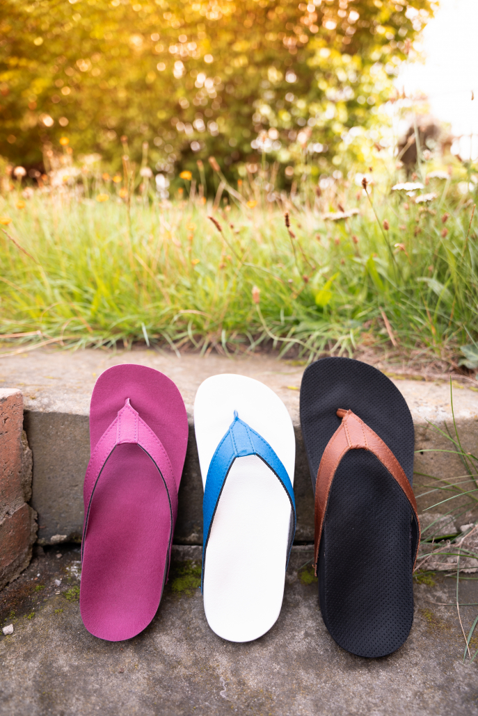 Riedel Flip Flops Auf dem Foto sieht man drei verschiedenfarbige Flip Flops, die auf einer Steintreppe angelehnt, an die zweite Stufe von oben stehen. Im Hintergrund sieht man Gras und den Ansatz von Büschen in der Unschärfe.