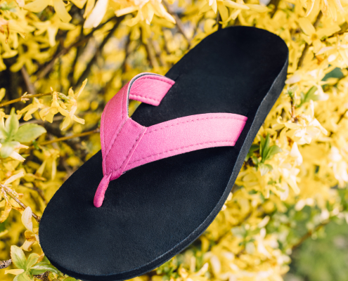 Auf dem Foto sieht man einen unserer individuellen Flip-Flops. Die Sohle ist schwarz, mit einem pinken Streifen in der Mitte und der Zehensteg ist ebenfalls pink. Der Flip-Flop steht in einem Busch mit gelben Blüten, um ein Frühlingsgefühl zu erwecken.
