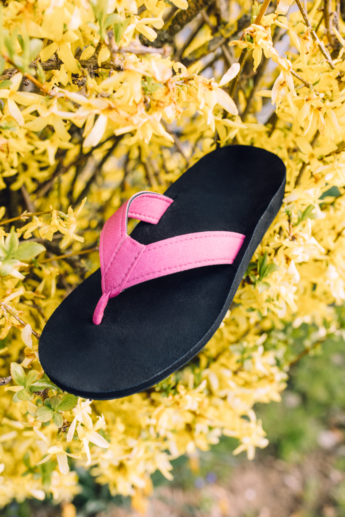 Auf dem Foto sieht man einen unserer individuellen Flip-Flops. Die Sohle ist schwarz, mit einem pinken Streifen in der Mitte und der Zehensteg ist ebenfalls pink. Der Flip-Flop steht in einem Busch mit gelben Blüten, um ein Frühlingsgefühl zu erwecken.