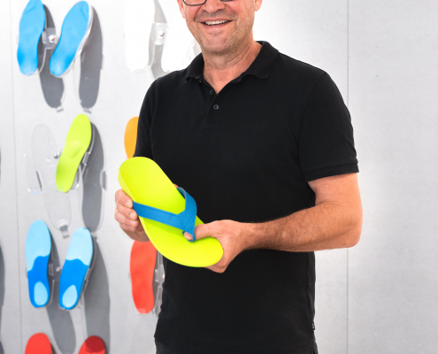 Flip-Flops, Sanitätshaus, Orthopädietechnik, Einlagen, Handwerk, Riedel Orthopädie, Gesundheit Auf dem Foto sieht man unseren Chef, Robert Riedel, wie er einen unserer Flip-Flops in der Hand hält und dabei lächelt. Der Flip-Flop hat die Farbe neongrün. Im Hintergrund sind Einlagen an einer Wand zu sehen.