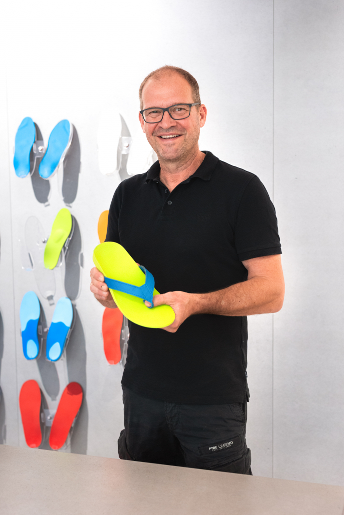 Auf dem Foto sieht man unseren Chef, Robert Riedel, wie er einen unserer Flip-Flops in der Hand hält und dabei lächelt. Der Flip-Flop hat die Farbe neongrün. Im Hintergrund sind Einlagen an einer Wand zu sehen.