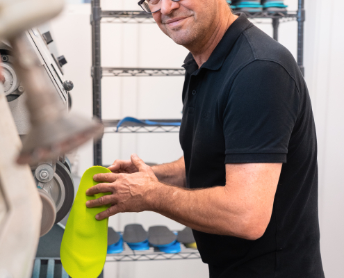 Flip-Flops, Sanitätshaus, Orthopädietechnik, Einlagen, Handwerk, Riedel Orthopädie, Gesundheit Auf dem Foto sieht man unseren Chef, Robert Riedel, an der Schleifmaschine, während er gerade an einem neongrünen Flip-Flop schleift. Im Hintergrund sieht man ein rollbares Gestell mit Einlagen.