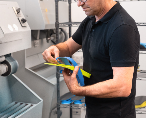 Flip-Flops, Sanitätshaus, Orthopädietechnik, Einlagen, Handwerk, Riedel Orthopädie, Gesundheit Auf dem Foto sieht man unseren Chef, Robert Riedel, an der Schleifmaschine stehend, während er gerade den Zehensteg in einen Flip-Flops einbaut. Im Hintergrund sieht man ein rollbares Gestell mit Einlagen.