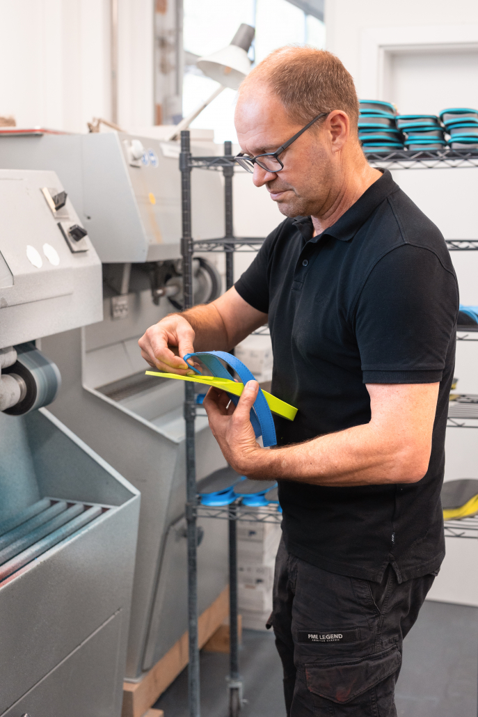 Auf dem Foto sieht man unseren Chef, Robert Riedel, an der Schleifmaschine stehend, während er gerade den Zehensteg in einen Flip-Flops einbaut. Im Hintergrund sieht man ein rollbares Gestell mit Einlagen.